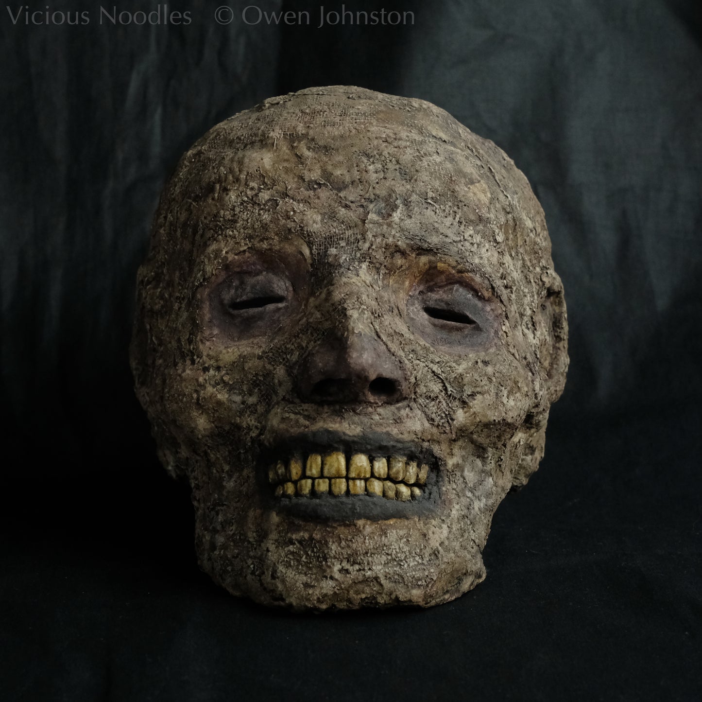 Replica mummified head, life size. It is well preserved, with nose and ears and a full set of yellowed teeth, and it is covered in bandages and fabric which appears highly aged and is painted various shades of brown. The skin features (eyes, nose, lips, ears) are painted deep brown.