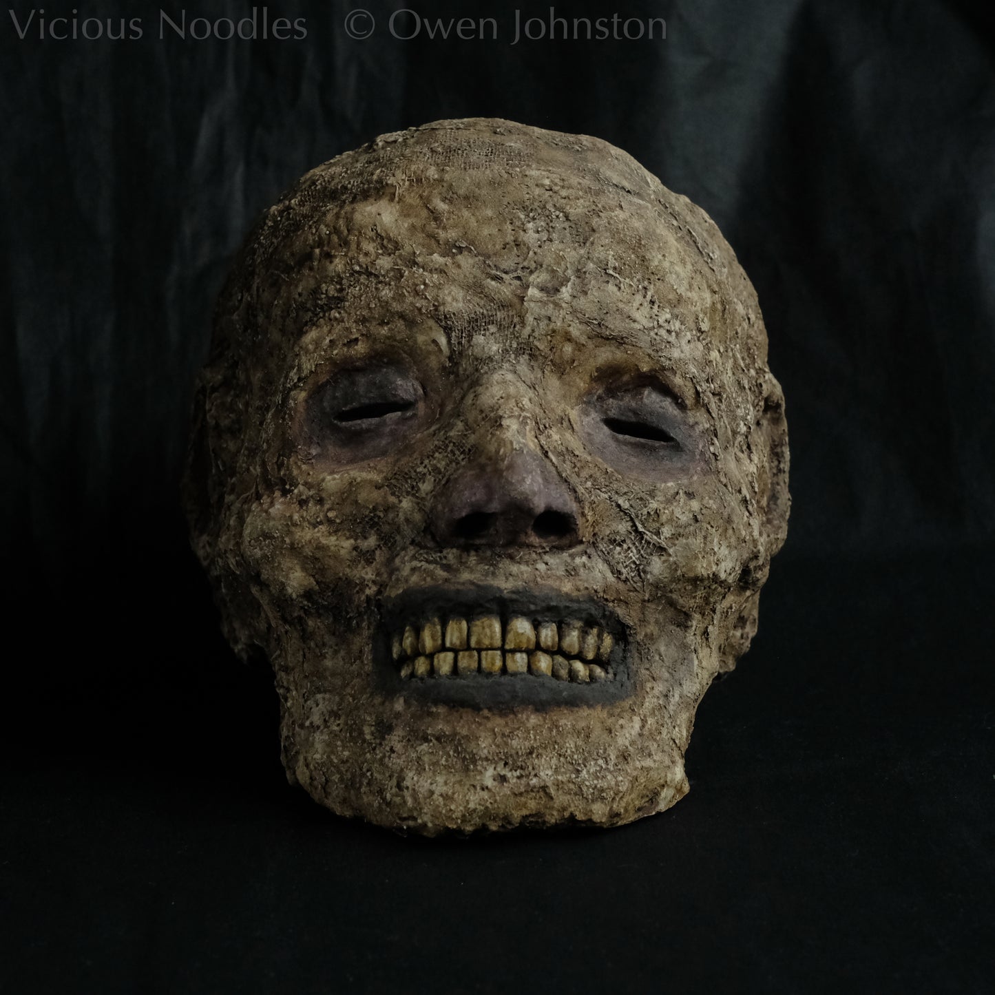 Replica mummified head, life size. It is well preserved, with nose and ears and a full set of yellowed teeth, and it is covered in bandages and fabric which appears highly aged and is painted various shades of brown. The skin features (eyes, nose, lips, ears) are painted deep brown.