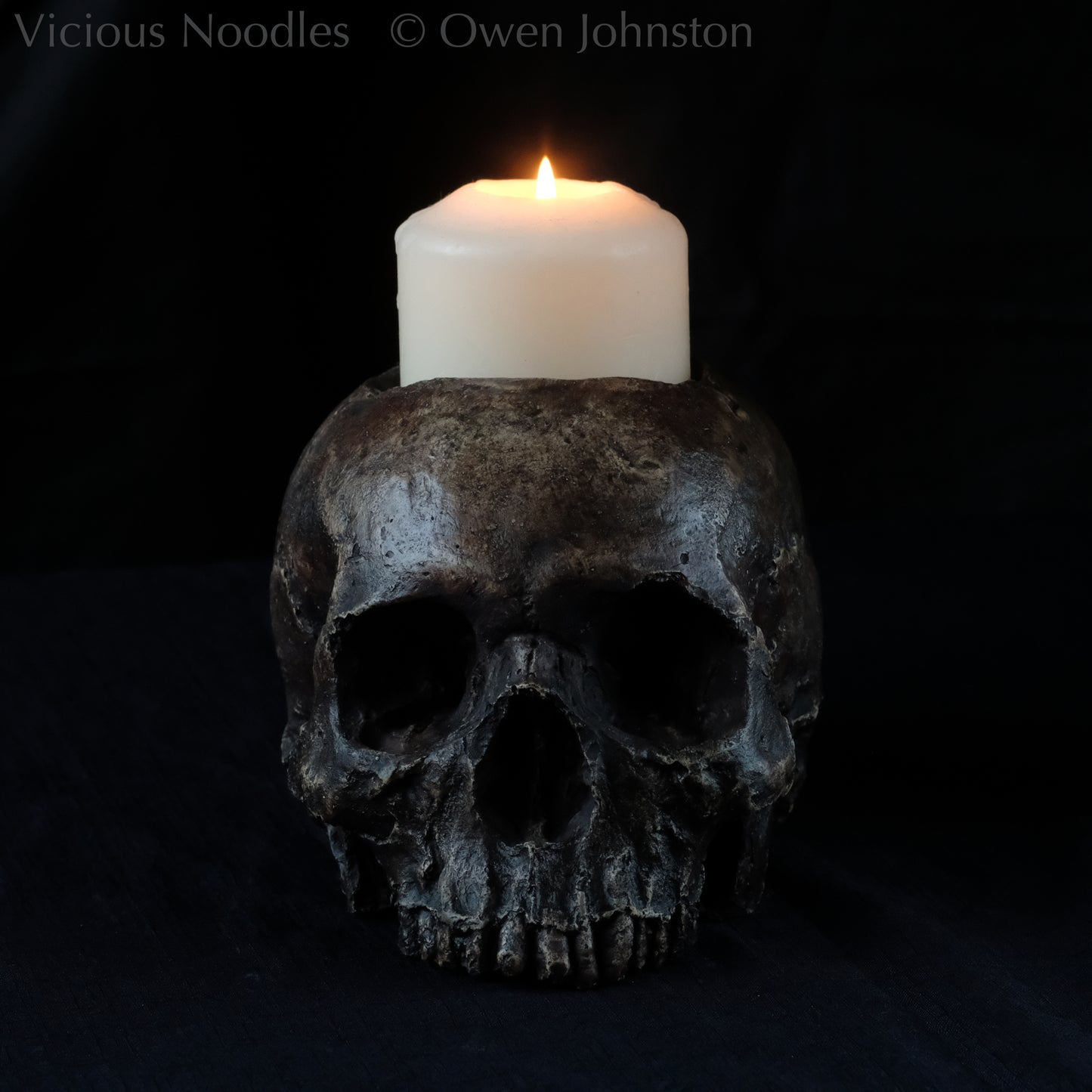 Human skull candle holder, without lower jaw, life size. It has a shallow hole on the top for a wide candle and is painted for a highly aged fossilised appearance with dark brown over lighter shades of brown. Lacquered for a polished finish.
