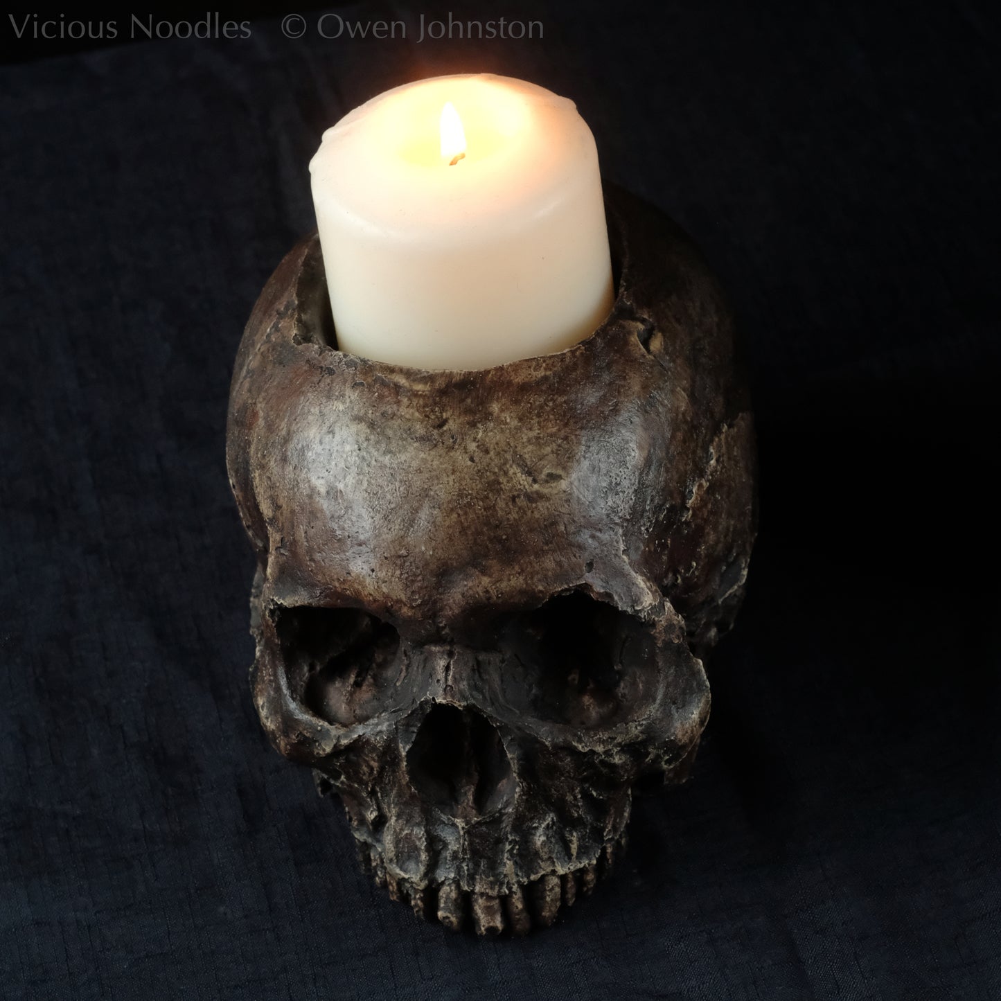 Human skull candle holder, without lower jaw, life size. It has a shallow hole on the top for a wide candle and is painted for a highly aged fossilised appearance with dark brown over lighter shades of brown. Lacquered for a polished finish.