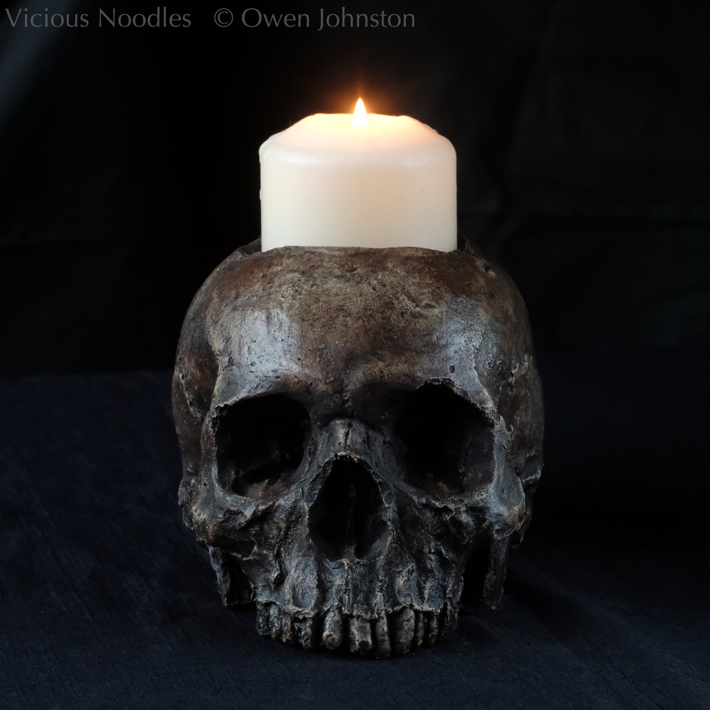 Human skull candle holder, without lower jaw, life size. It has a shallow hole on the top for a wide candle and is painted for a highly aged fossilised appearance with dark brown over lighter shades of brown. Lacquered for a polished finish.