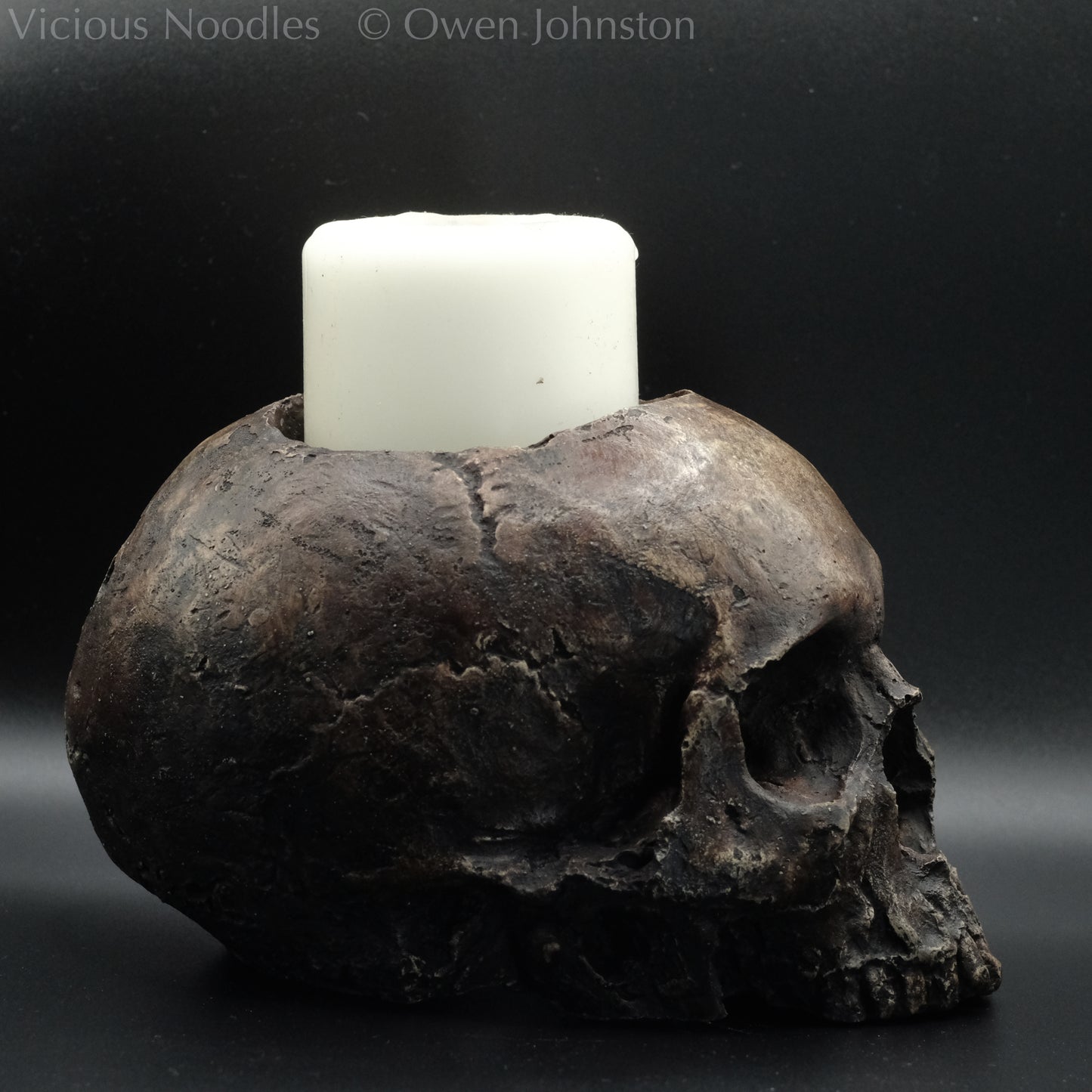 Human skull candle holder, without lower jaw, life size. It has a shallow hole on the top for a wide candle and is painted for a highly aged fossilised appearance with dark brown over lighter shades of brown. Lacquered for a polished finish.