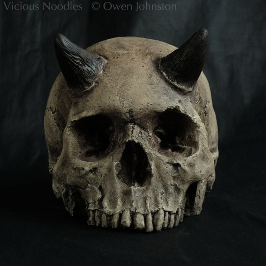 Horned human skull replica without jaw, life size, warm ivory bone colour with dark weathering, finished with lacquer.
