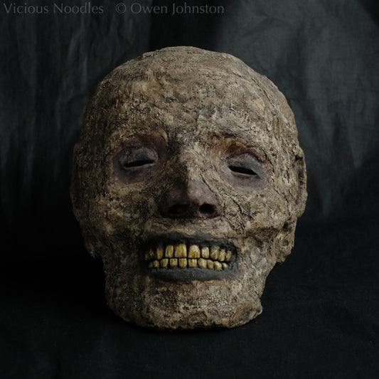 Replica mummified head, life size. It is well preserved, with nose and ears and a full set of yellowed teeth, and it is covered in bandages and fabric which appears highly aged and is painted various shades of brown. The skin features (eyes, nose, lips, ears) are painted deep brown.
