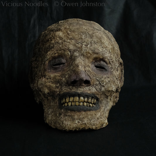 Replica mummified head, life size. It is well preserved, with nose and ears and a full set of yellowed teeth, and it is covered in bandages and fabric which appears highly aged and is painted various shades of brown. The skin features (eyes, nose, lips, ears) are painted deep brown.
