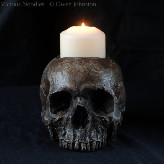 Human skull candle holder, without lower jaw, life size. It has a shallow hole on the top for a wide candle and is painted for a highly aged fossilised appearance with dark brown over lighter shades of brown. Lacquered for a polished finish.